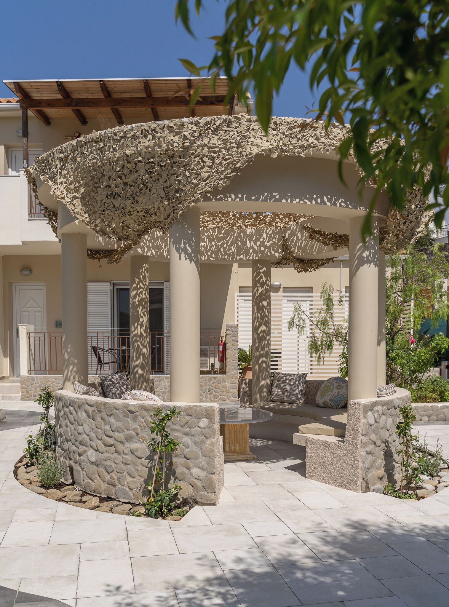 Ein überdachter Außenbereich mit Steinsäulen und einer Pergola, umgeben von einer Steinmauer und Topfpflanzen.