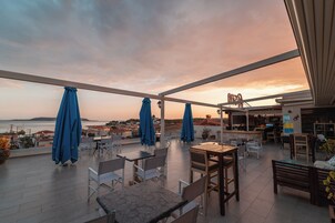 Una terraza con mesas y sillas, sombrillas azules y vistas al mar y edificios.