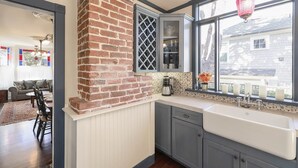 A kitchen with a brick wall, a large window with a hanging lantern, and a view of a house outside. - 3812 Little House - Historic Home, Walk to the Bay (Pacific Grove)
