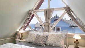 A bedroom with a bed, two bedside tables, a window with curtains, and a view of houses. - 3812 Little House - Historic Home, Walk to the Bay (Pacific Grove)