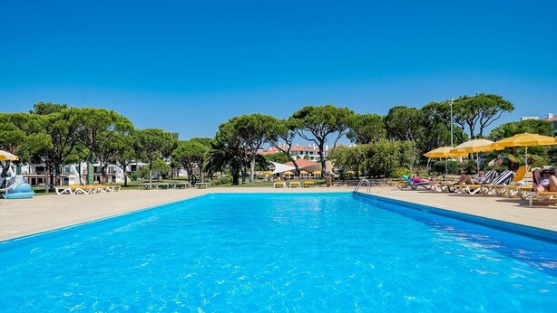 A swimming pool with lounge chairs and umbrellas, surrounded by trees and buildings. - Vila Sol: Nice apartment with large south-facing terrace and view of own park (Quarteira)