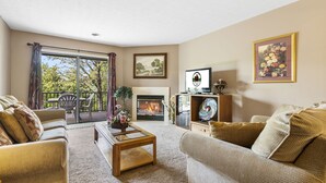 Una sala de estar con chimenea, televisor y un balcón con vistas a árboles.