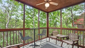 A covered outdoor deck with wooden furniture, a ceiling fan, and a view of trees.