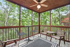Une terrasse extérieure couverte, dotée de meubles en bois, d’un ventilateur de plafond et offrant une vue sur les arbres.