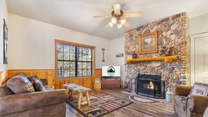 A cozy living room with a stone fireplace, wooden furniture, and a large window.