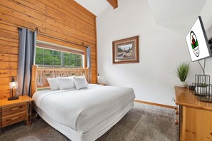Une chambre à coucher avec une tête de lit en bois, un lit recouvert de draps blancs, une table de chevet avec une lampe, une fenêtre avec des rideaux, un tableau encadré accroché au mur et un téléviseur posé sur un meuble en bois.
