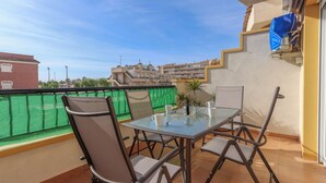 A balcony with a glass table and chairs, a green mesh fence, and a view of buildings in the background. - 032 - Parque y Playa 001 - comfortHOLIDAYS (Santa Pola)