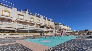 A playground with swings and a slide, surrounded by a tiled area and buildings with balconies. - 032 - Parque y Playa 001 - comfortHOLIDAYS (Santa Pola)