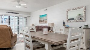 A dining area with a wooden table and white chairs, a ceiling fan, and a beach-themed painting on the wall. - Fully remodeled 2 bedroom condo on the ocean block in North OC (Ocean City)