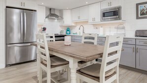 A modern kitchen with white cabinetry, stainless steel appliances, and a wooden dining table. - Fully remodeled 2 bedroom condo on the ocean block in North OC (Ocean City)