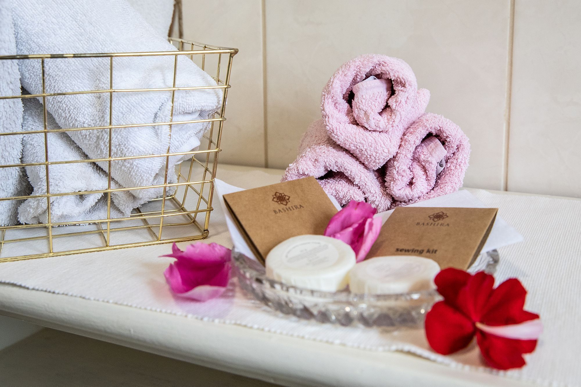 Une salle de bain bien rangée, avec un panier en fil d’or contenant des serviettes blanches, une serviette rose et un plateau en verre avec une trousse de couture et des fleurs roses.