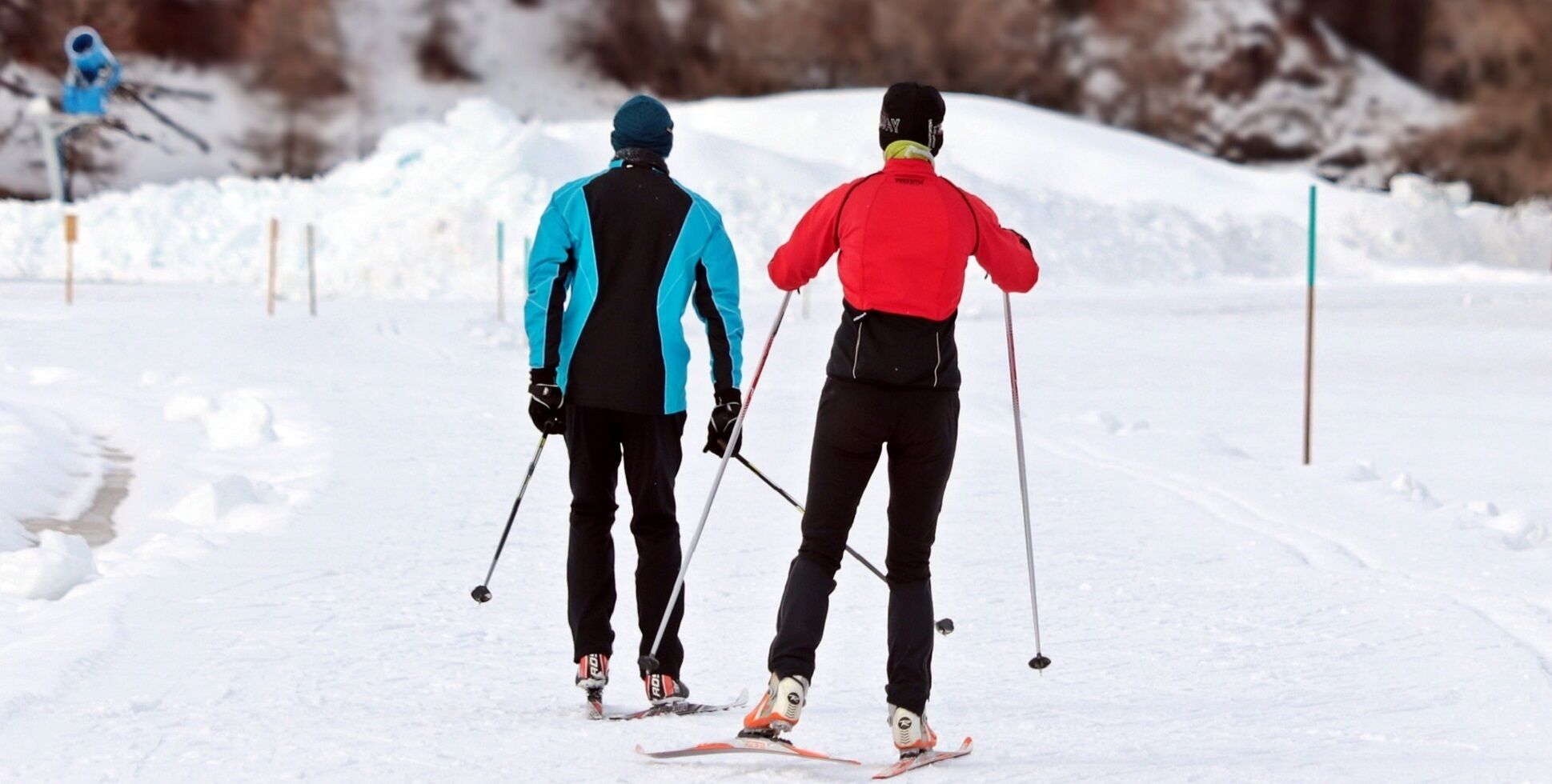 Dos personas esquiando por un sendero nevado, usando bastones de esquí, con chaquetas y cascos.