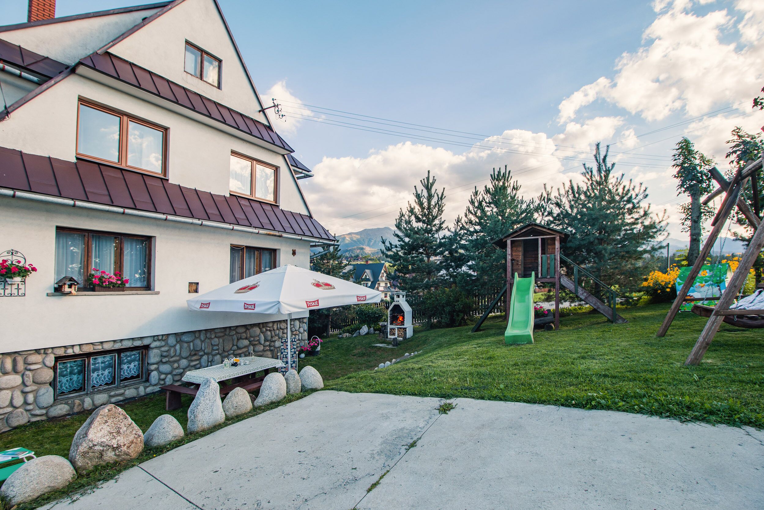 Ein Haus mit steinernem Fundament, rotem Dach und einer Terrasse mit Tisch und Stühlen.