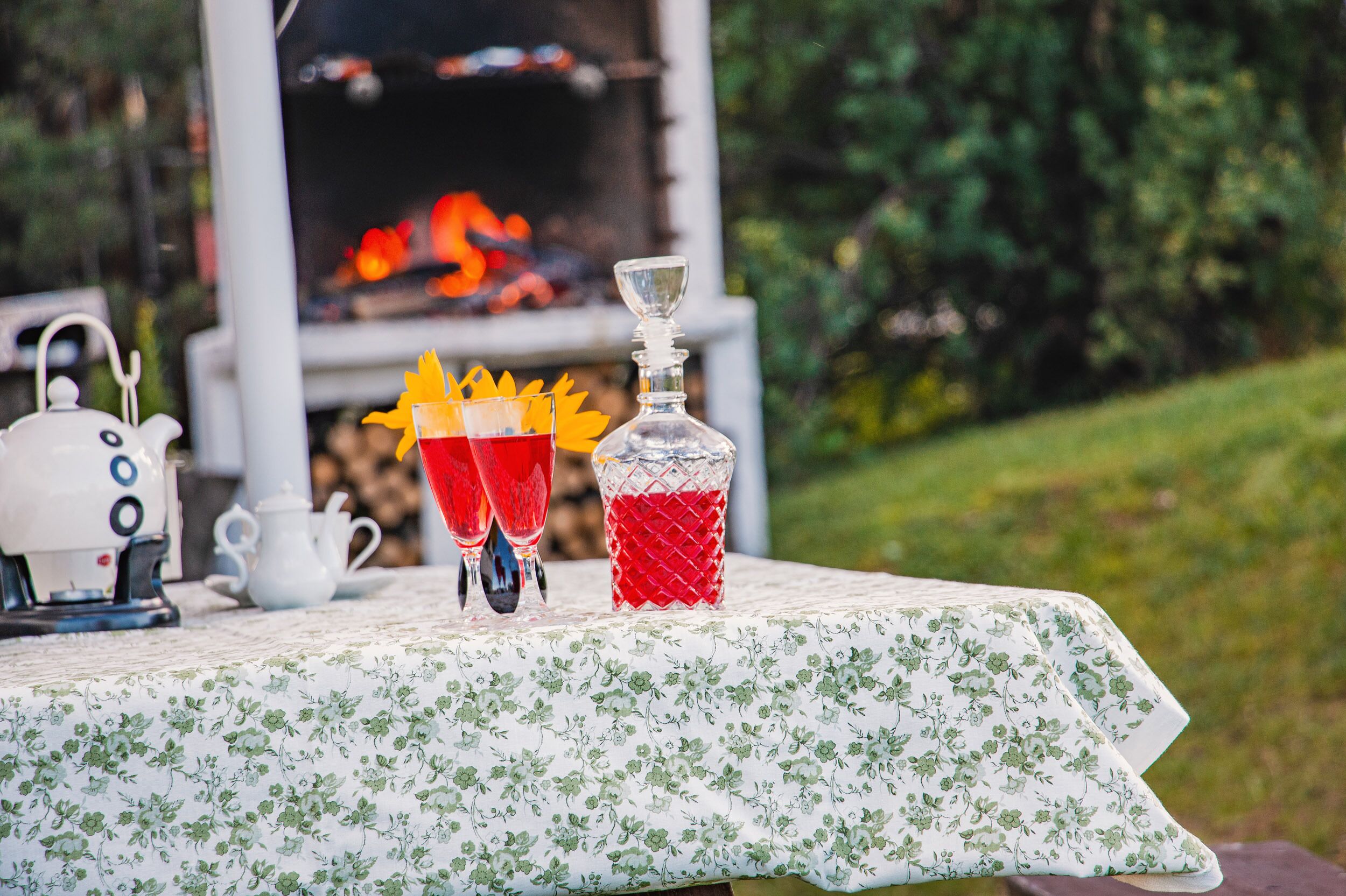 A table with a floral tablecloth, a teapot, two glasses with a red drink, and a decanter with a red liquid.