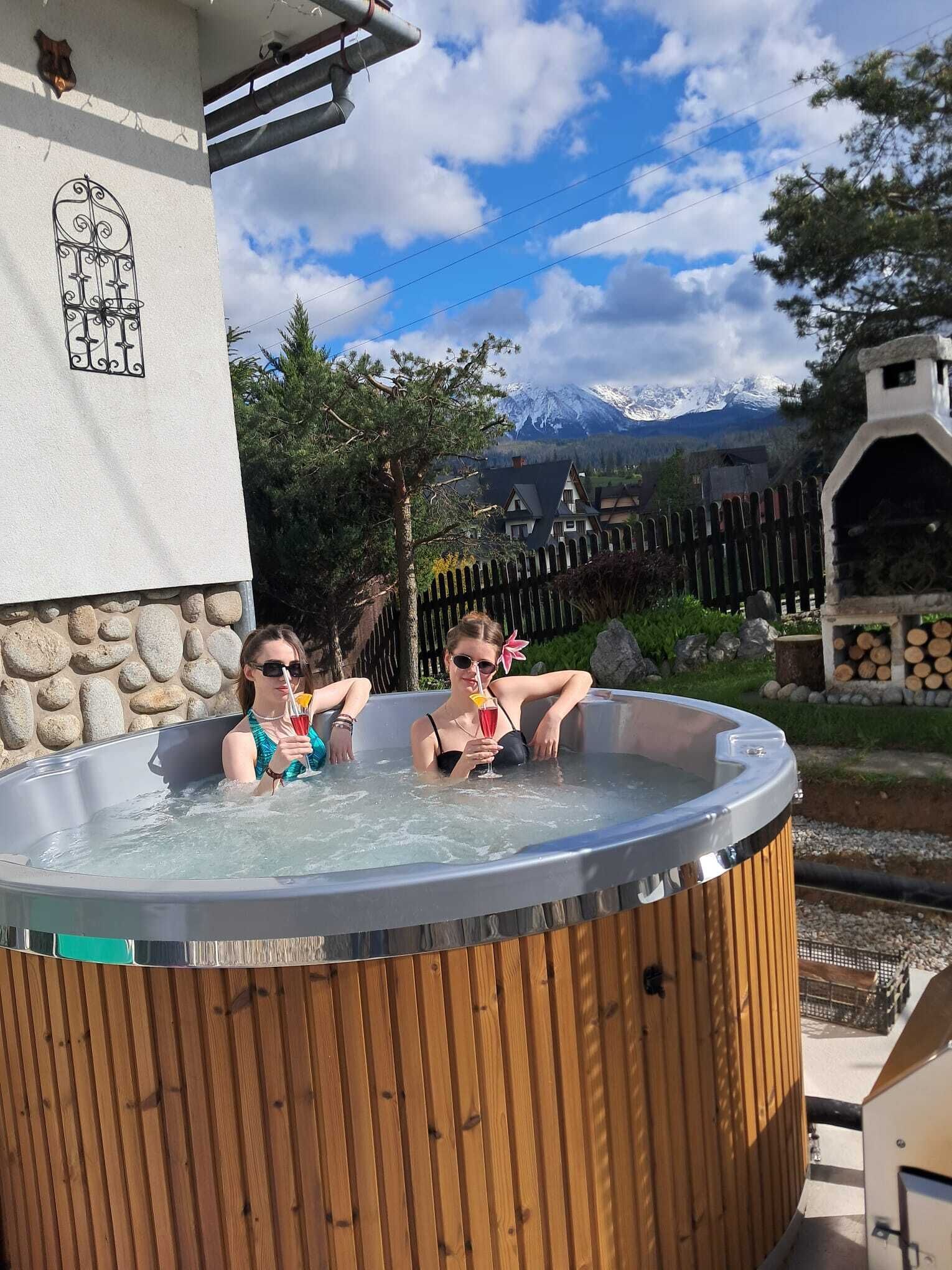 Dos personas en un jacuzzi de madera con vista a una montaña al fondo.
