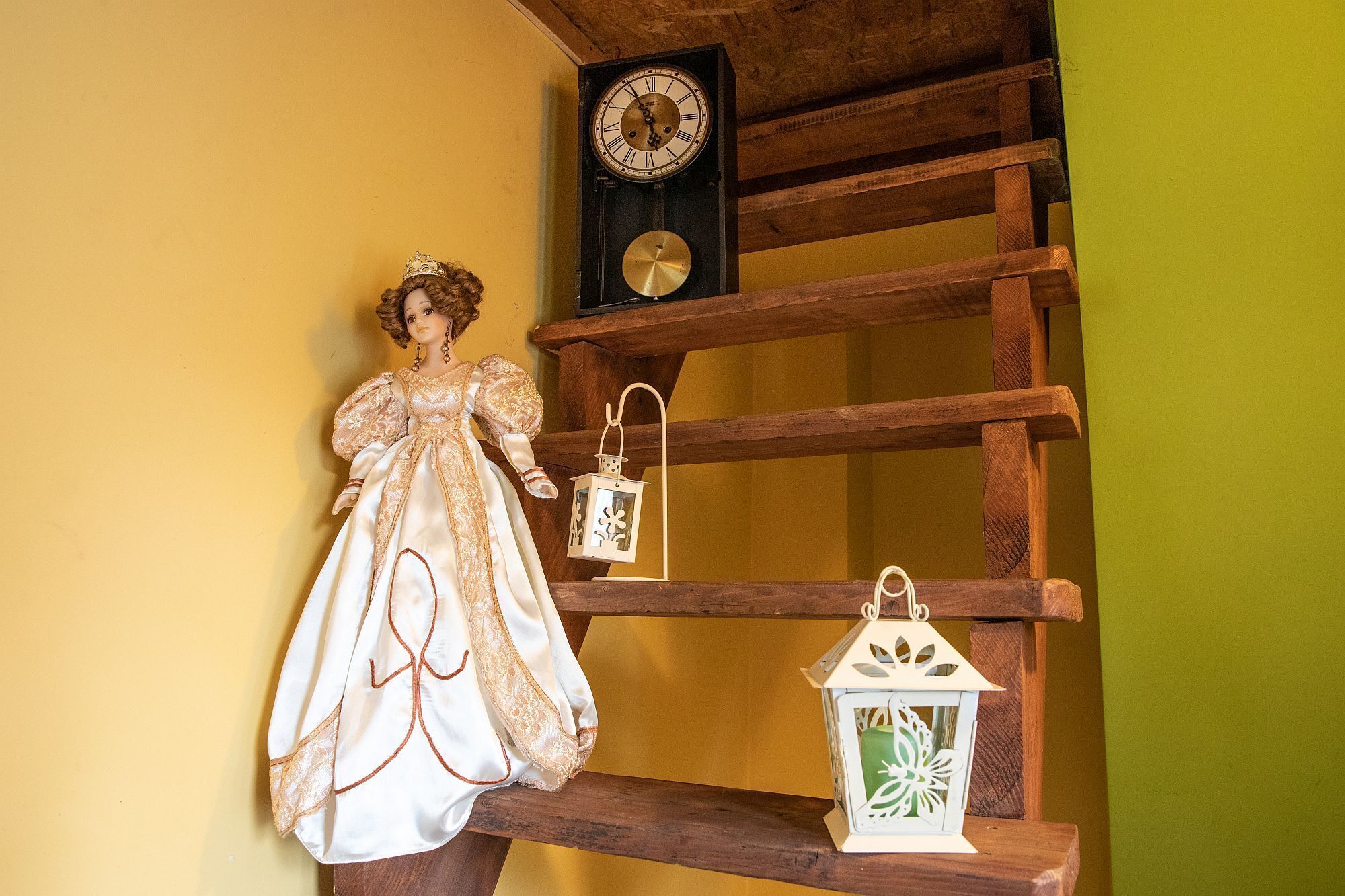 A doll in a vintage dress on a wooden shelf with two white lanterns and a clock.