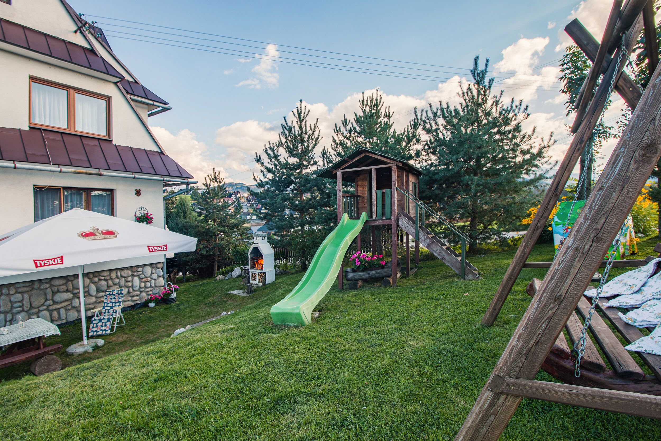 A house with a stone foundation, a white canopy, and a wooden playhouse with a slide.