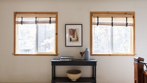 Una habitación con dos ventanas, una consola de madera oscura con un cuenco, un cuadro enmarcado y un jarrón con flores.