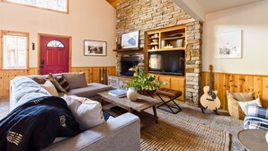 A cozy living room with a stone fireplace, wooden beams, a red door, and a television mounted on the wall. - Two-Cabin Buyout + 2 Hot Tubs | Walk to Snow Summit | Jumper & Jackhorn (Big Bear Lake)