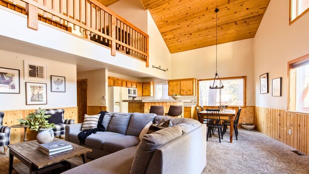 A spacious living area with a wooden ceiling, a dining table with chairs, and a large grey sectional sofa. - Two-Cabin Buyout + 2 Hot Tubs | Walk to Snow Summit | Jumper & Jackhorn (Big Bear Lake)