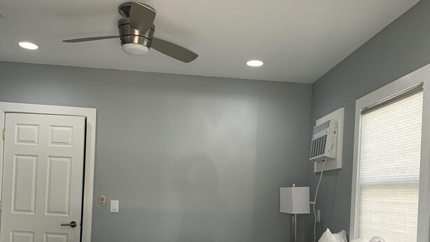 Une chambre à coucher comprenant un lit, un ventilateur de plafond, des spots encastrés, une porte, une table de chevet avec une lampe et une fenêtre avec des stores.