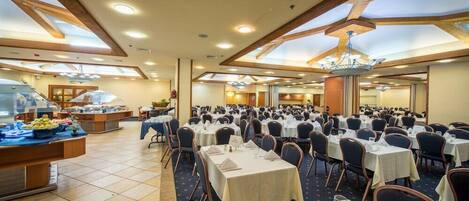 A large banquet hall with numerous round tables, a buffet area, and a high ceiling with skylights.