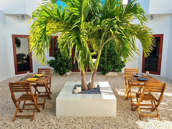 A patio area with two wooden tables, each surrounded by four chairs, set on a gravel surface. A large tree with broad leaves is centrally located, and there are potted plants and a small decorative item on the tables.