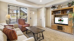 Una sala de estar con sofá, sillón y mesa de centro. Hay un televisor sobre un mueble de madera y una ventana con persianas.