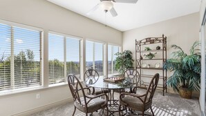 A dining area with a glass table and metal chairs, large windows with blinds, a ceiling fan, and a plant shelf.