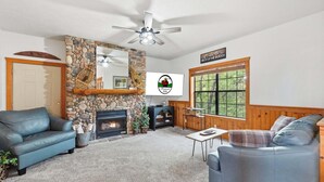 A cozy living room with a stone fireplace, a ceiling fan, and a large window with a view of trees. - Walk-in Log Cabin with Fireplace & Screened-in Balcony - Heart of Branson (Branson)