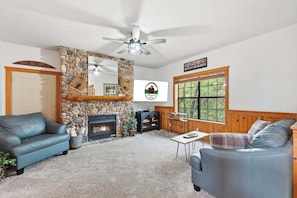 Un salon chaleureux avec une cheminée en pierre, un ventilateur de plafond et une grande fenêtre donnant sur les arbres.