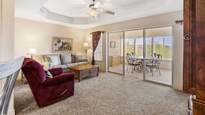 A spacious living room with a red velvet chair, a wooden coffee table, a sofa, and a painting on the wall. There is a sliding glass door leading to an outdoor patio with a dining table and chairs. - 1 Bedroom Walk-in Golf Condo feat. Sunroom with a Beautiful Property View (Branson)