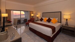 A hotel room with a large bed, two bedside tables with lamps, a desk with a computer and glasses, a chair, and a view of the ocean. - Tesoro Manzanillo (Manzanillo)