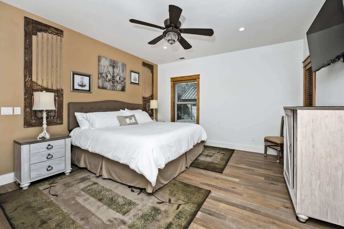 Ein Schlafzimmer mit Bett, Deckenventilator, Schminktisch, Fenster und einem an der Wand montierten Fernseher.