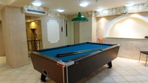 Une table de billard dans une pièce dont le sol est recouvert de carreaux, avec une lampe verte suspendue au plafond et une chaise.