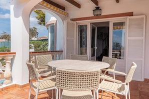 A covered patio with a round table and wicker chairs, a sliding glass door, and a view of the surrounding area. - V105 | Villa S'Illot (Torre Soli Nou)