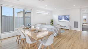 Une pièce moderne et bien éclairée, avec une table à manger en bois, des chaises blanches, un canapé et un téléviseur fixé au mur.
