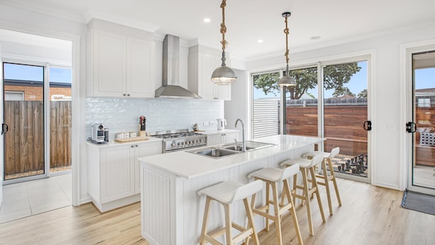 Une cuisine moderne avec des meubles blancs, un îlot central doté de tabourets de bar, une cuisinière à gaz et une grande fenêtre donnant sur un jardin.
