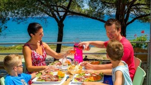 Une famille est assise à une table dressée à l’extérieur, prête à prendre son repas, avec une vue sur un plan d’eau.