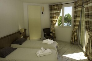 A hotel room with two beds, a chair, a window with curtains, and a view of greenery outside. - Village des Iles (Taglio-Isolaccio)