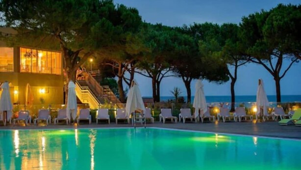 Un espace aménagé au bord de la piscine avec des chaises longues et des parasols, un bâtiment doté d’un escalier et des arbres.