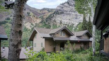 A two-story house with a sloped roof, surrounded by greenery and mountains in the background.