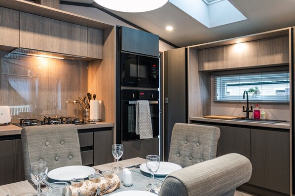 A modern RV kitchen with a dining area, equipped with a stove, oven, microwave, and a window with blinds. - Luxury Lodge in Stunning Surroundings (York)