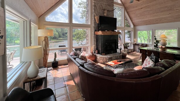 A spacious living room with a stone fireplace, leather sofa, and large windows overlooking a lake. - Camelot on Lake Hamilton | Dock, Hot Tub + Theater (Hot Springs)