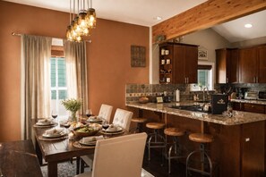 A dining area with a wooden table set for six, bar stools, a kitchen with wooden cabinets, and a hanging light fixture.