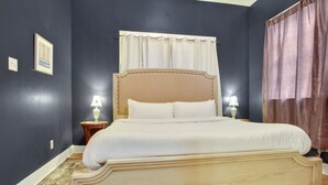 Una cama con estructura de madera y ropa de cama blanca, dos lámparas de noche, un cuadro en la pared y una ventana con cortinas.