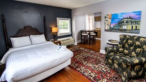 Une chambre à coucher avec un grand lit, une chaise, une table avec des tabourets, un tableau et une fenêtre avec des stores.
