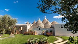 A traditional stone house with conical roofs, a well-maintained garden with potted plants, and a clear blue sky.