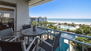 A balcony with a table and chairs overlooking a beach and a pool area.