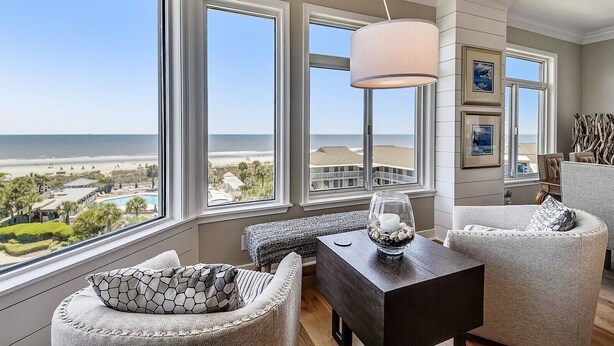 A room with large windows overlooking a beach, a dark wooden table, and a comfortable seating area.